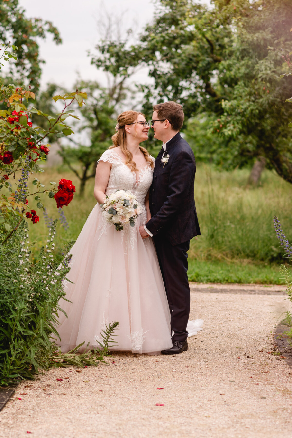 christian-lorenz.net Eine Braut im weißen Kleid und ein Bräutigam im dunklen Anzug stehen dicht beieinander auf einem Gartenweg, umgeben von Grün und roten Blumen.