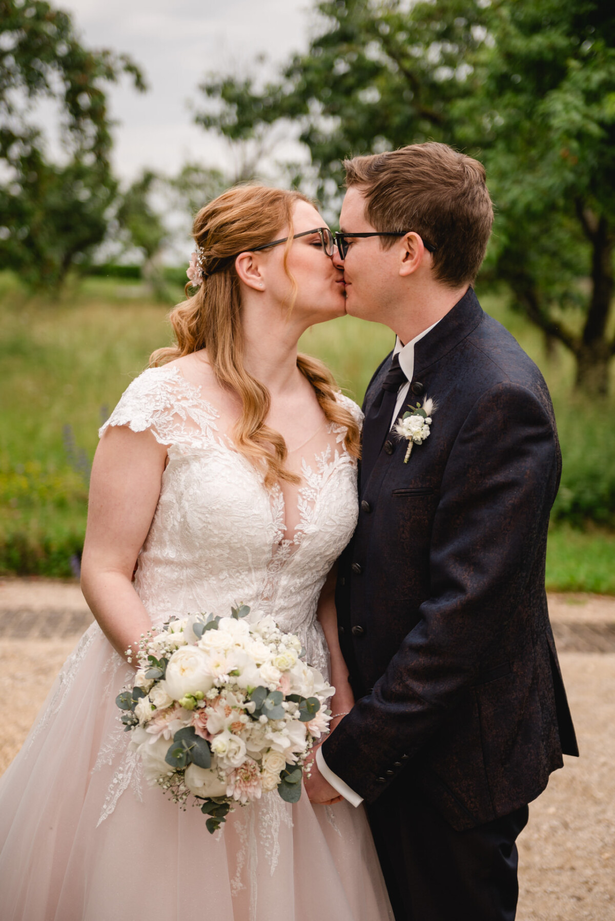 Braut und Bräutigam küssen sich im Freien. Die Braut hält einen weißen Blumenstrauß in der Hand und trägt ein Spitzenkleid. Der Bräutigam trägt eine Brille und einen dunklen Anzug. Im Hintergrund sind Bäume und ein Weg zu sehen.