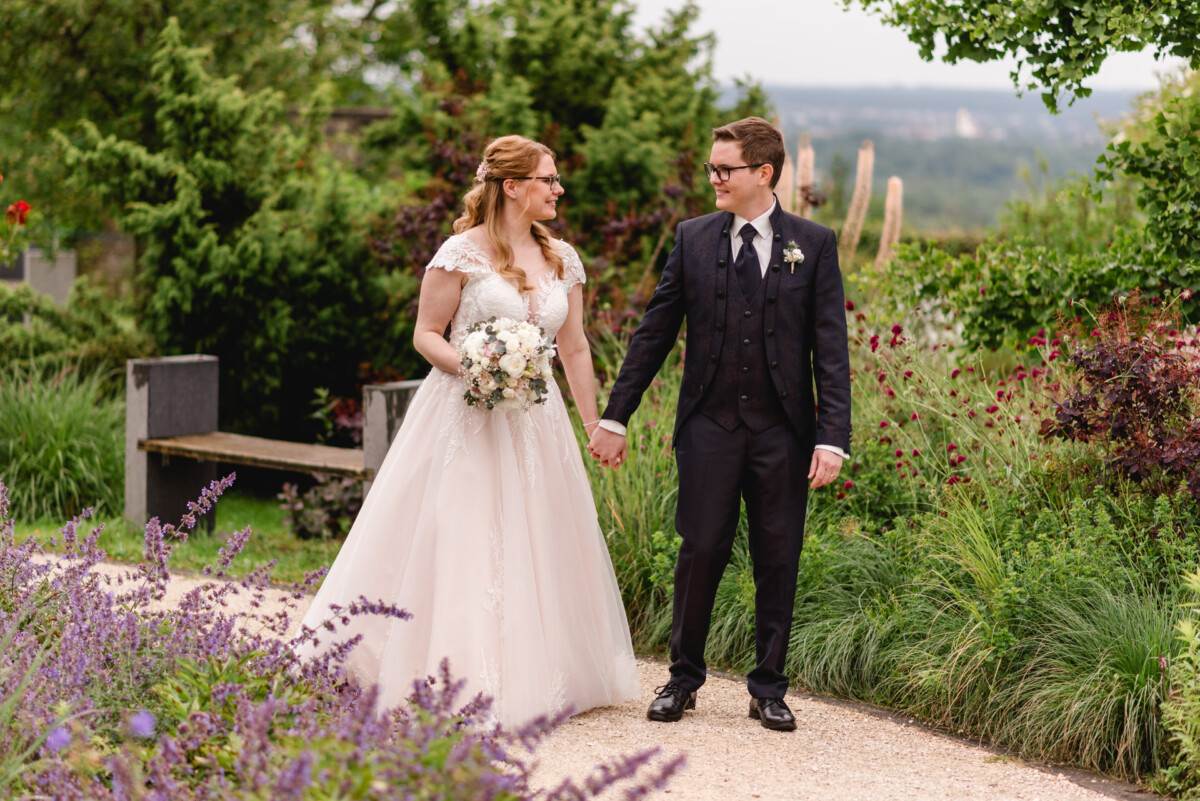christian-lorenz.net Braut im weißen Kleid und Bräutigam im dunklen Anzug halten Händchen und lächeln in einer Gartenkulisse mit Blumen und Grün.