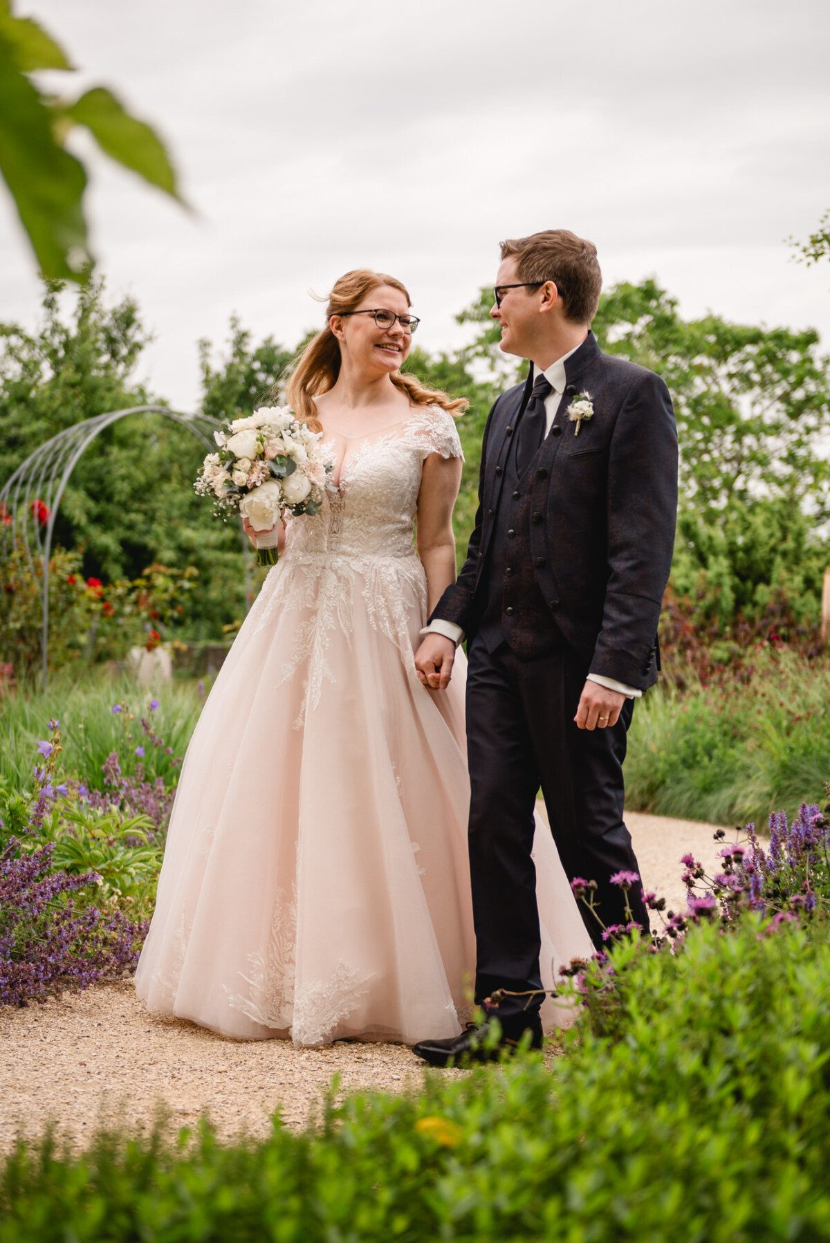 Braut und Bräutigam gehen Hand in Hand einen Gartenweg entlang. Die Braut hält einen Blumenstrauß, während der Bräutigam Anzug und Ansteckblume trägt. Üppiges Grün umgibt sie.