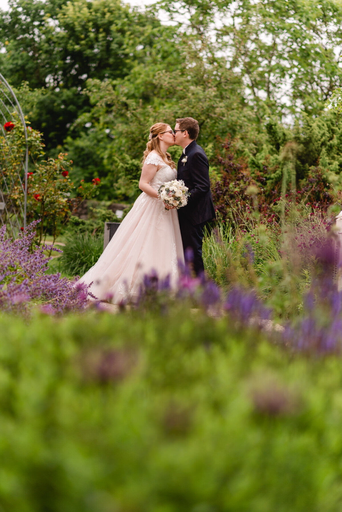 christian-lorenz.net Ein Paar in Hochzeitskleidung küsst sich in einem Garten. Die Frau trägt ein weißes Kleid und hält einen Blumenstrauß in der Hand, der Mann einen dunklen Anzug. Blumen und Grün umgeben sie.