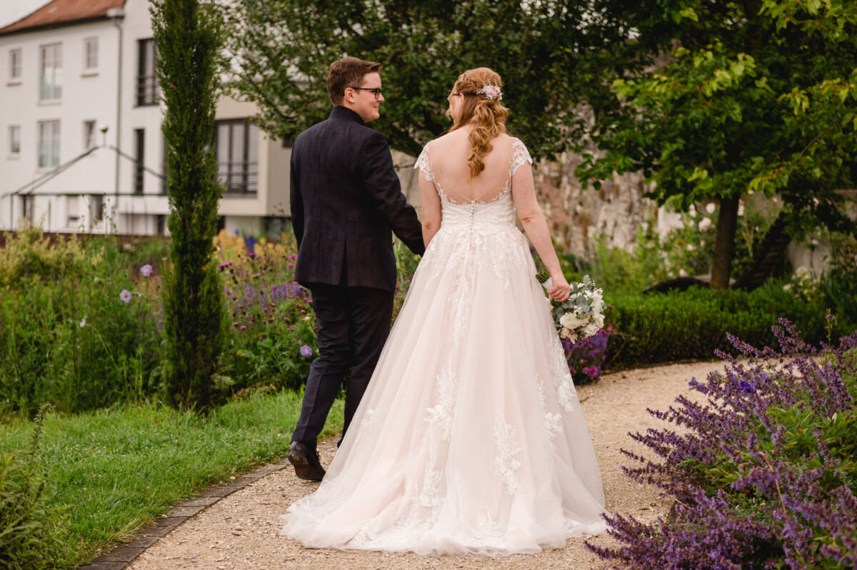 Eine Braut und ein Bräutigam gehen Hand in Hand einen Gartenweg entlang. Die Braut trägt ein weißes Kleid und hält einen Blumenstrauß in der Hand, umgeben von Grün und lila Blumen.