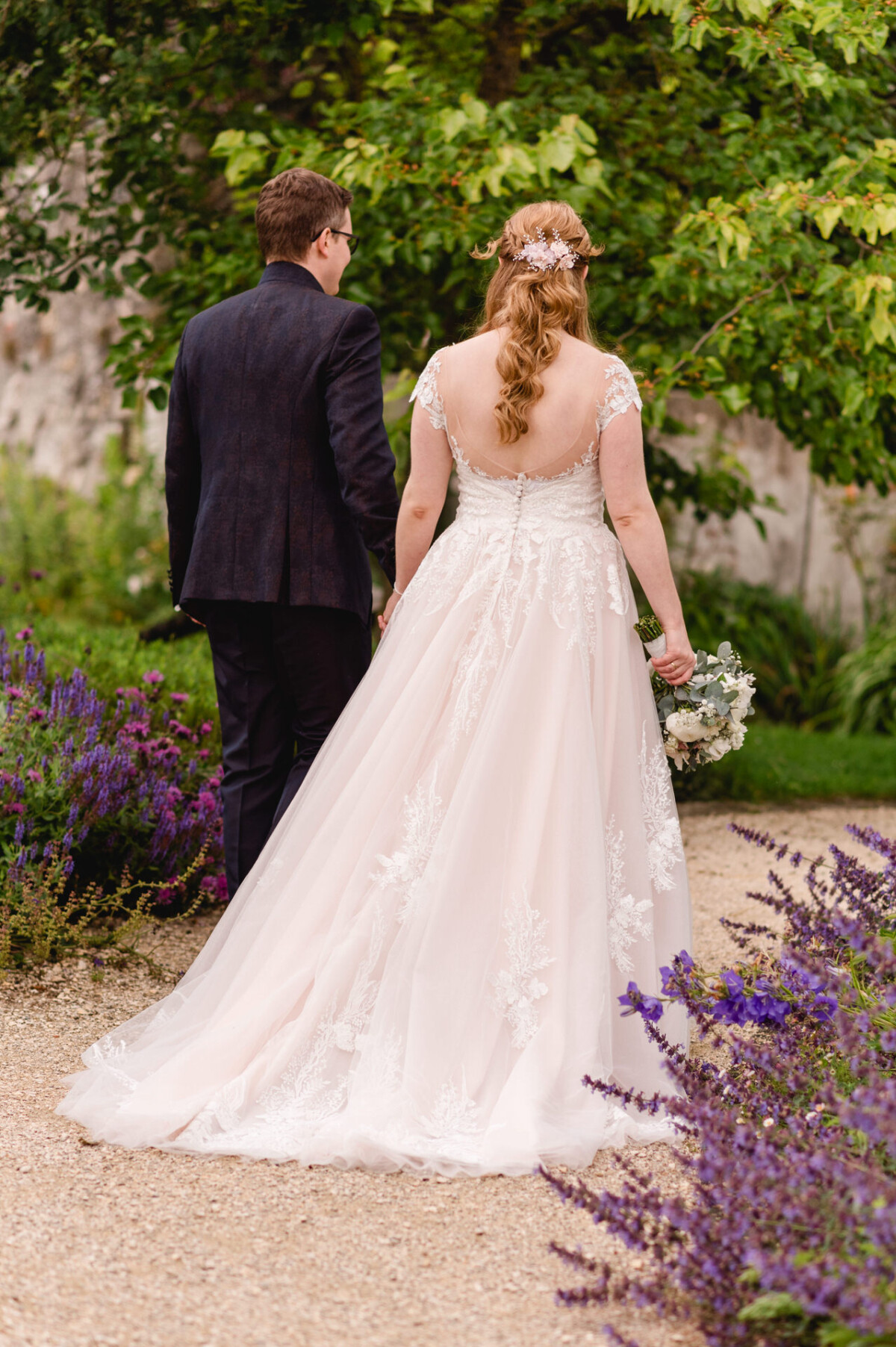 christian-lorenz.net Braut und Bräutigam gehen auf einem Gartenweg, umgeben von grünem Laub und lila Blumen. Das Spitzenkleid der Braut ist mit floralen Details und einem Blumenstrauß verziert. Der Bräutigam trägt einen dunklen Anzug.