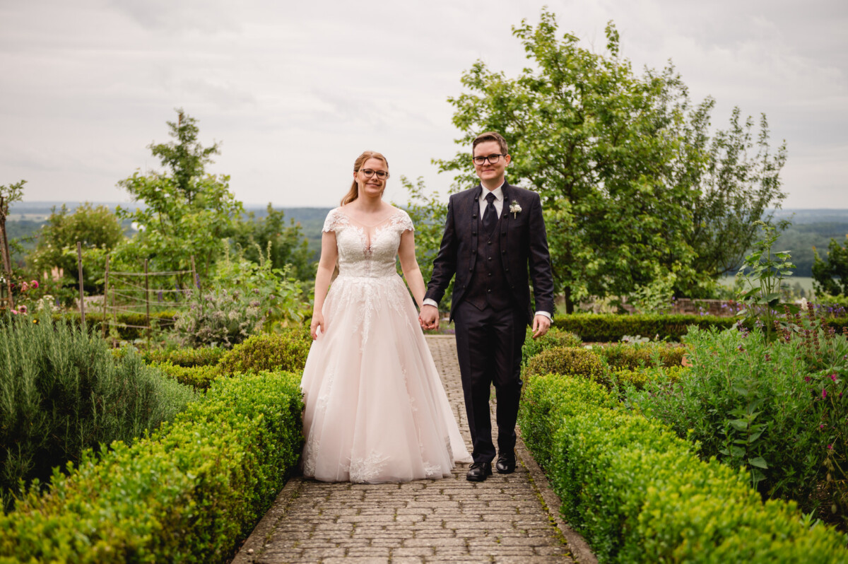 Eine Braut im weißen Kleid und ein Bräutigam im dunklen Anzug gehen Hand in Hand einen Gartenweg entlang, umgeben von Grün.