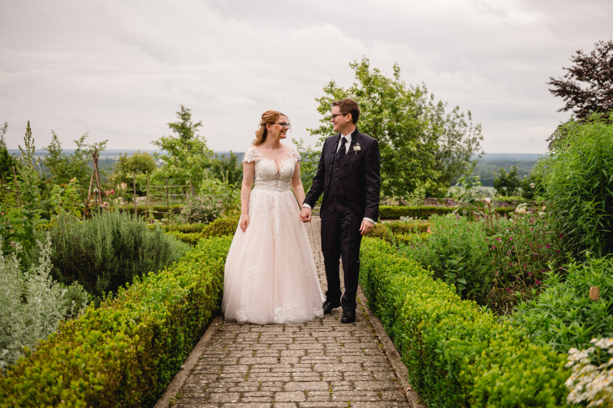 Ein Paar in Hochzeitskleidung geht Hand in Hand auf einem Steinpfad, umgeben von Grün und einem bewölkten Himmel.