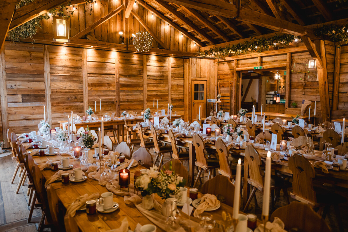 Rustikaler Bankettsaal aus Holz mit langen Tischen, geschmückt mit Blumenschmuck, Kerzen und Gedecken, bereit für eine Veranstaltung. Deckenleuchten tauchen den Raum in ein warmes Licht.