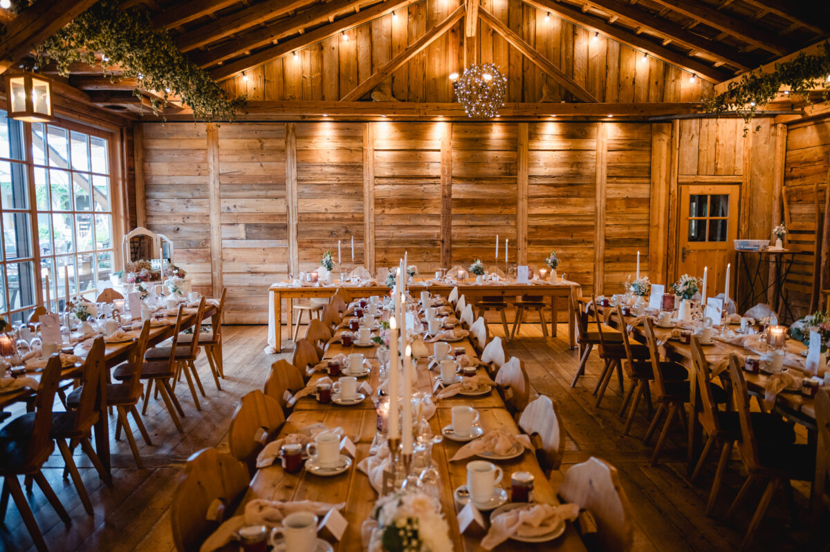 Rustikaler Bankettsaal aus Holz mit langen, gedeckten Tischen für eine Veranstaltung. Die Dekoration umfasst weiße Kerzen, Grünpflanzen, Geschirr und kleine Blumengestecke unter warmer Deckenbeleuchtung.