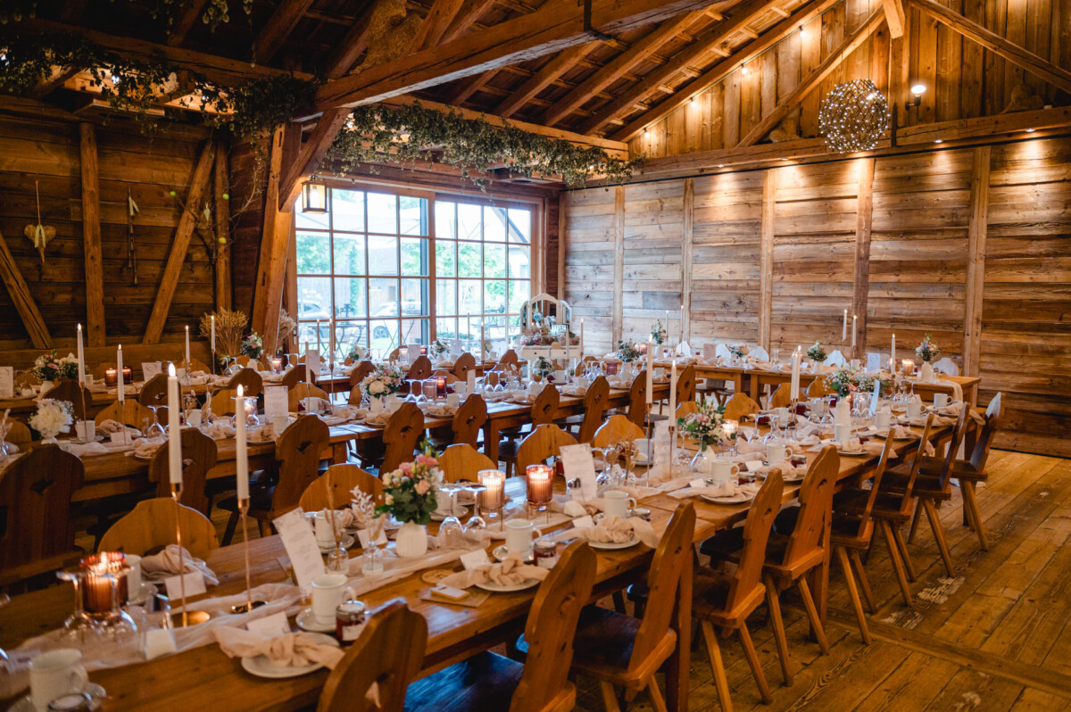 Rustikales Interieur mit Holzwänden und -decke, langen Holztischen mit Kerzen, Geschirr und Blumengestecken zum Essen und großen Fenstern, die natürliches Licht hereinlassen.