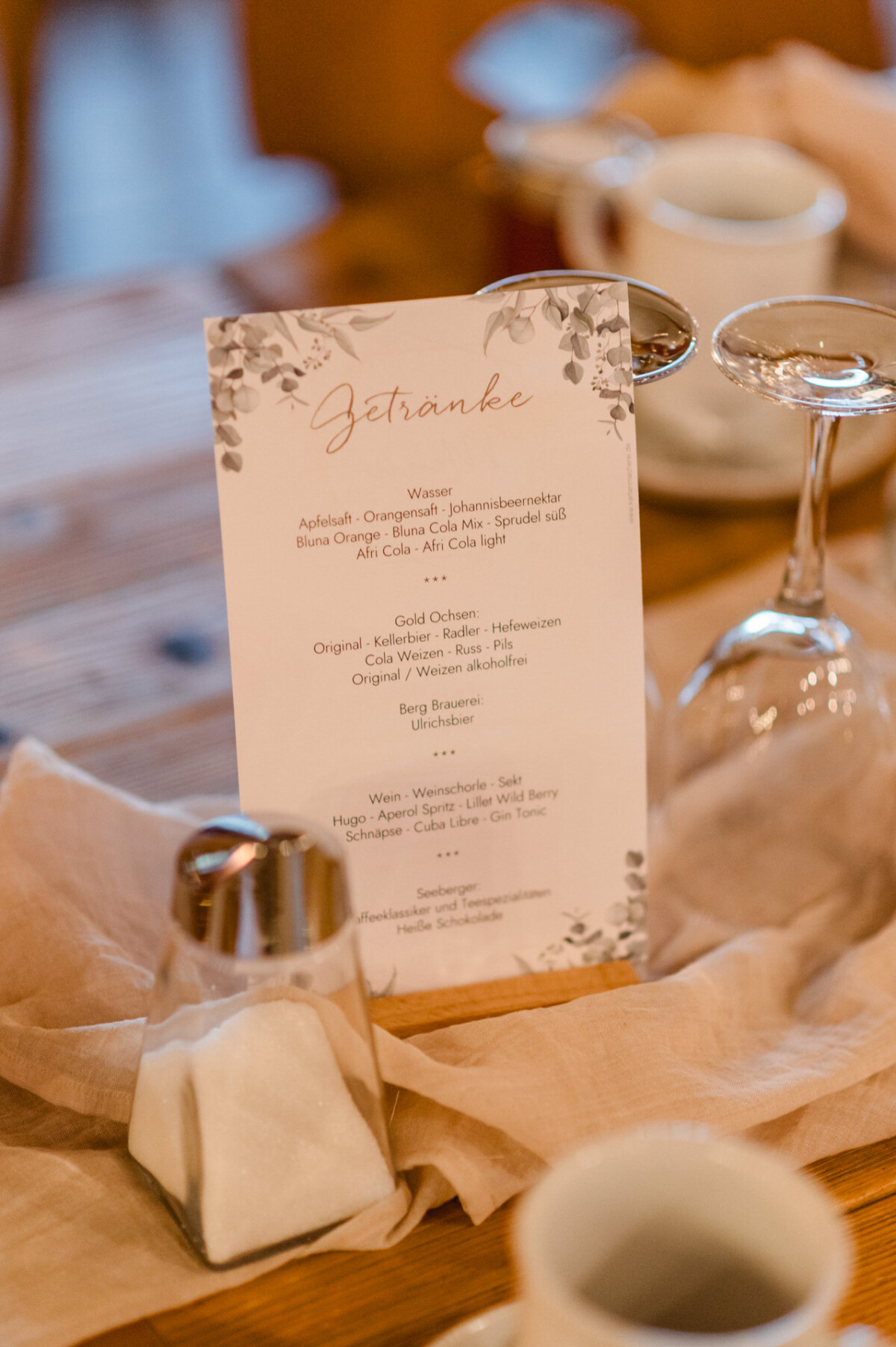 christian-lorenz.net Eine Speisekarte mit der Überschrift „Getränke“ auf einem Holztisch, umgeben von einem Salzstreuer, einem umgedrehten Glas und einem weißen Becher, darunter ein beiges Tuch.