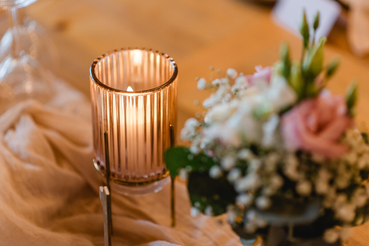 christian-lorenz.net Eine brennende Kerze in einem gerippten Glashalter neben einem kleinen Strauß rosa und weißer Blumen auf einem mit beigem Stoff gedeckten Tisch.
