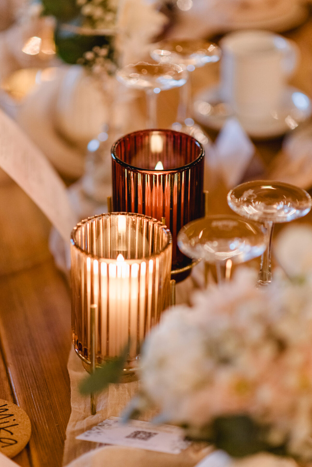 christian-lorenz.net Brennende Kerzen in gerippten Glashaltern sind auf einem Holztisch von weißen Blumen und elegantem Glasgeschirr umgeben.