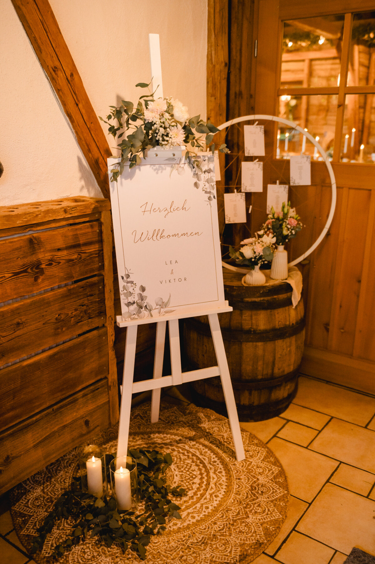 Hochzeits-Willkommensschild auf Staffelei mit Blumenschmuck, neben einem Holzfass, umgeben von Kerzen, in einem rustikalen Raum.