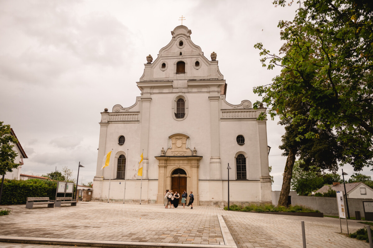 Vorderansicht einer Kirche im Barockstil mit weißer Fassade, Bogenfenstern und drei runden Öffnungen. Eine Gruppe von Menschen steht davor, im Hintergrund Bäume und ein bewölkter Himmel.