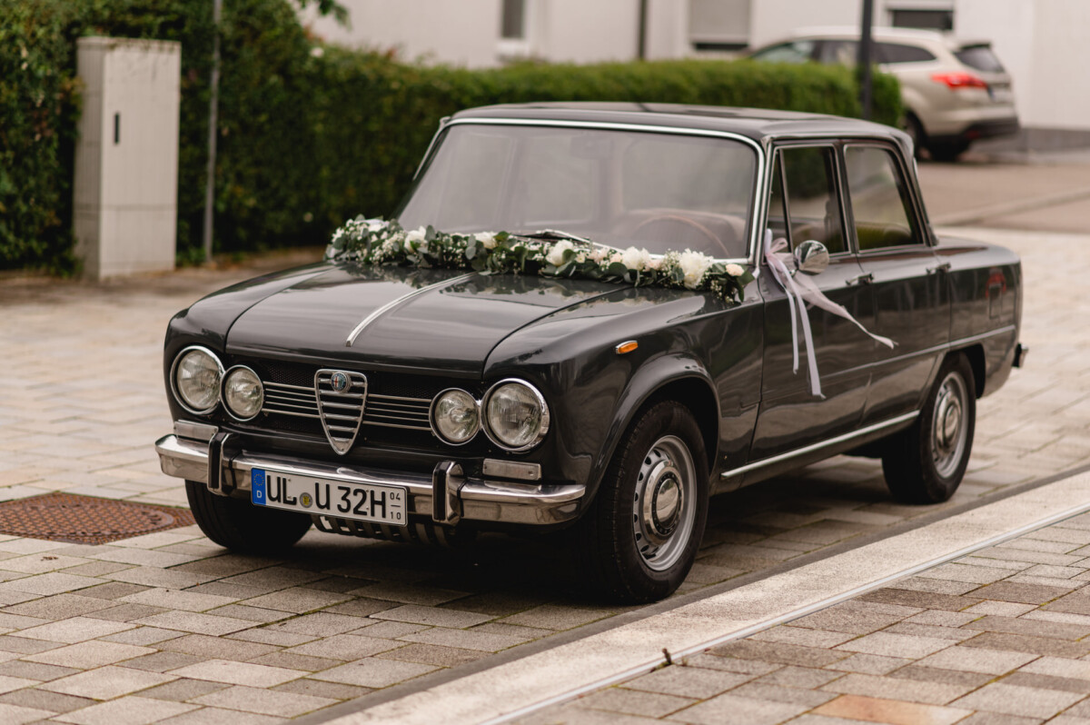 christian-lorenz.net Ein klassisches schwarzes Auto mit weißen Blumen auf der Motorhaube steht auf einer Kopfsteinpflasterstraße.