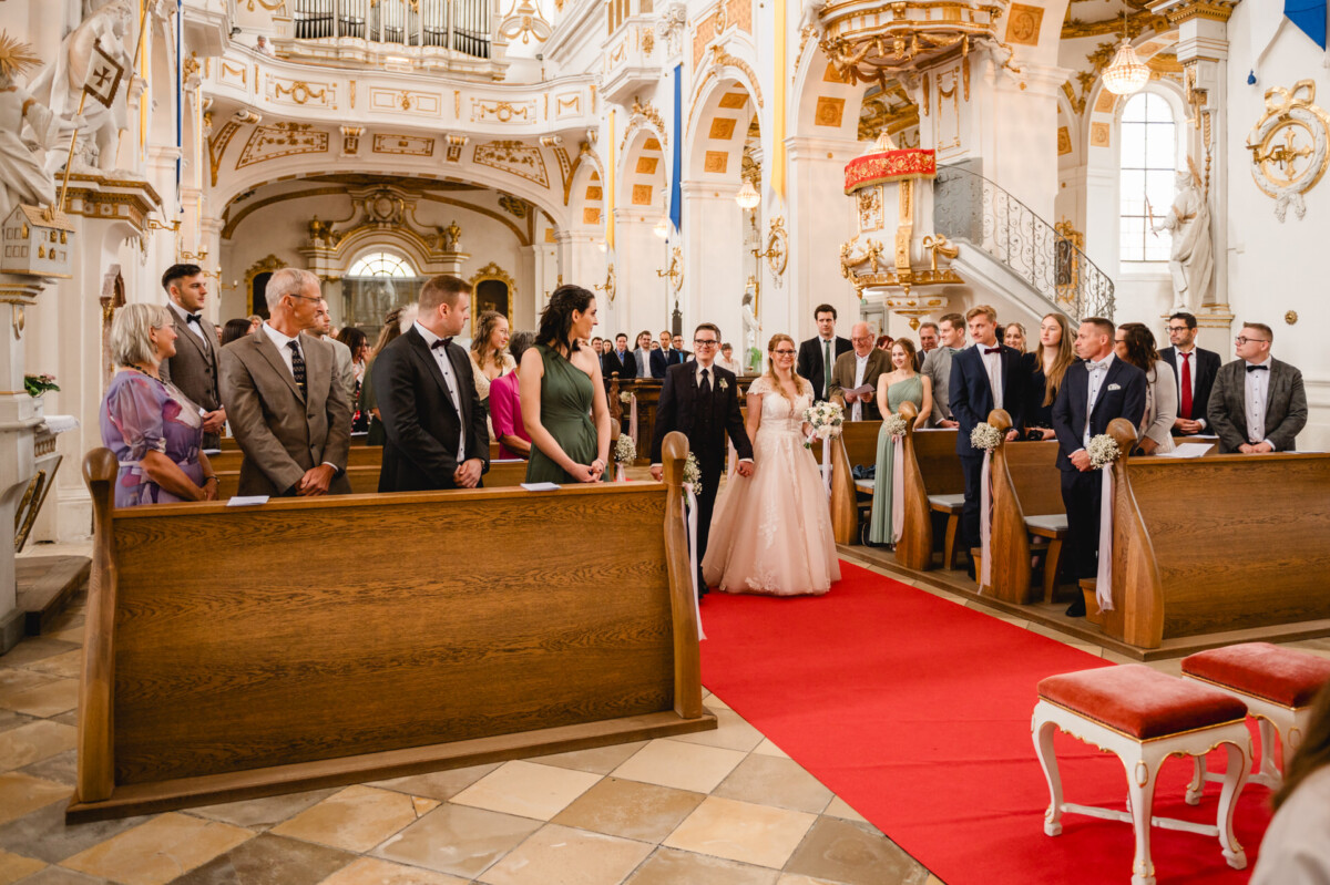 Eine Hochzeitszeremonie in einer geschmückten Kirche. Braut und Bräutigam stehen am Altar und blicken auf eine Gruppe sitzender Gäste. Die Kirchenbänke sind mit weißen Blumen geschmückt.
