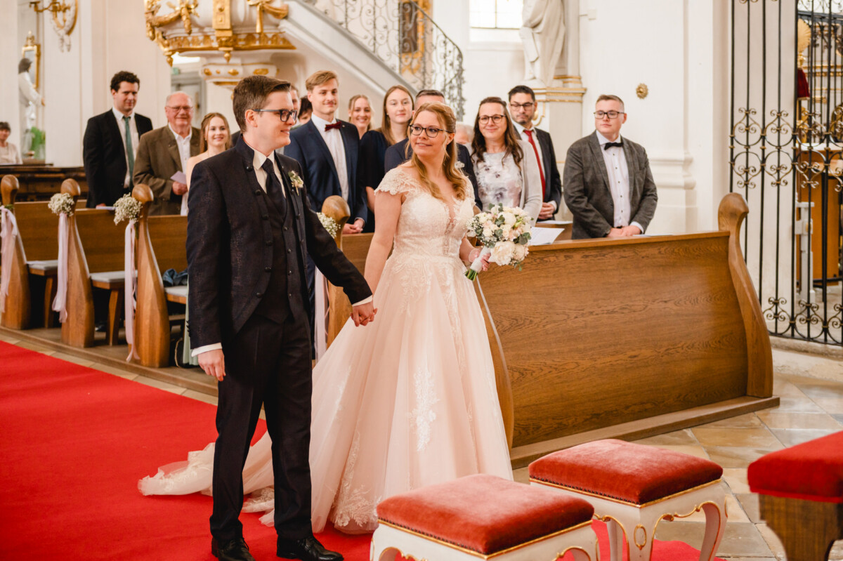 Braut und Bräutigam stehen in einer Kirche händchenhaltend am Altar. Hinter ihnen sitzen Gäste auf Holzbänken. Die Braut hält einen Blumenstrauß in der Hand und trägt ein weißes Kleid; der Bräutigam trägt einen dunklen Anzug.