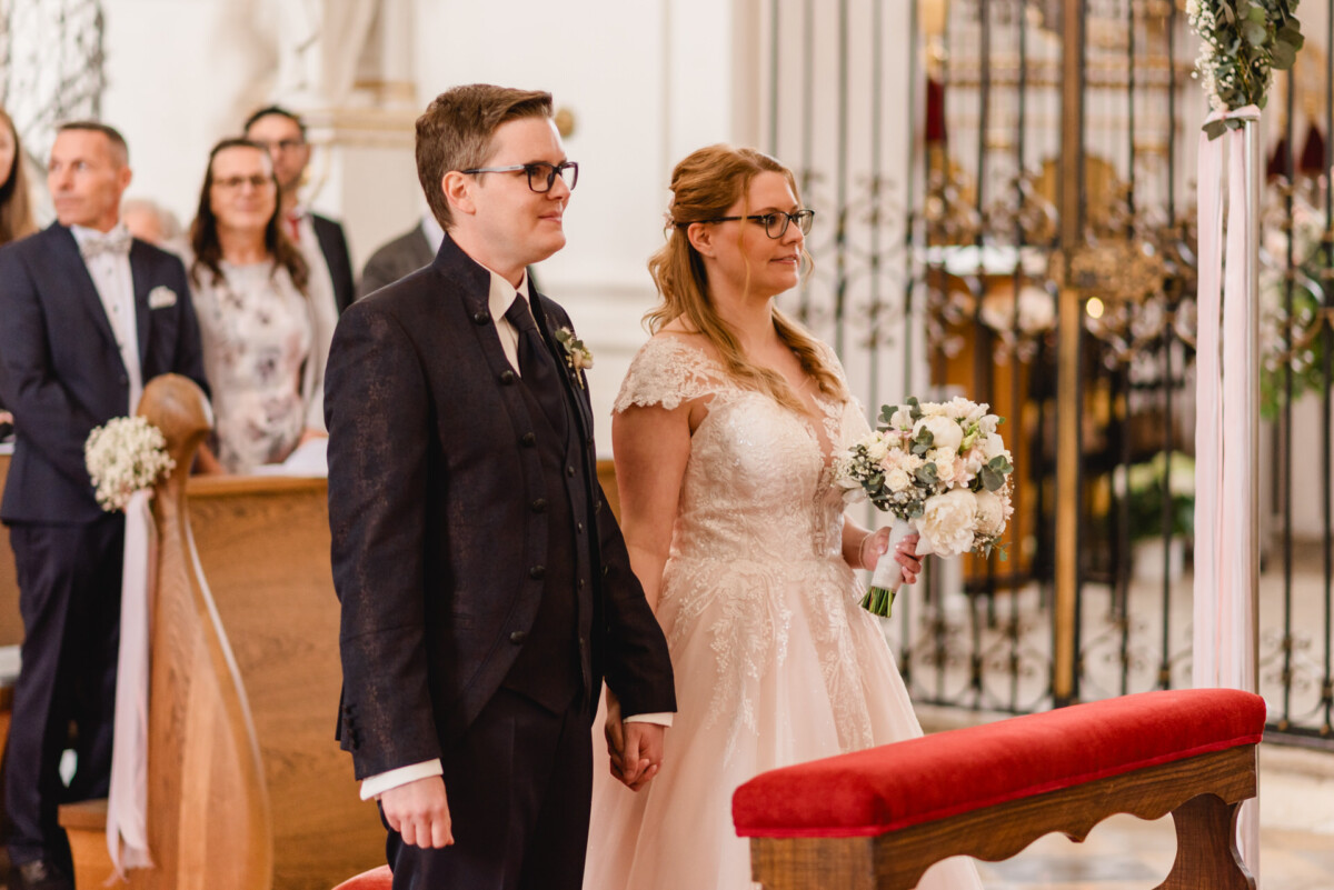 christian-lorenz.net Braut und Bräutigam stehen während einer Hochzeitszeremonie gemeinsam am Altar. Beide halten sich an den Händen und blicken nach vorne. Die Braut hält einen Blumenstrauß in der Hand. Im Hintergrund sitzen die Gäste.
