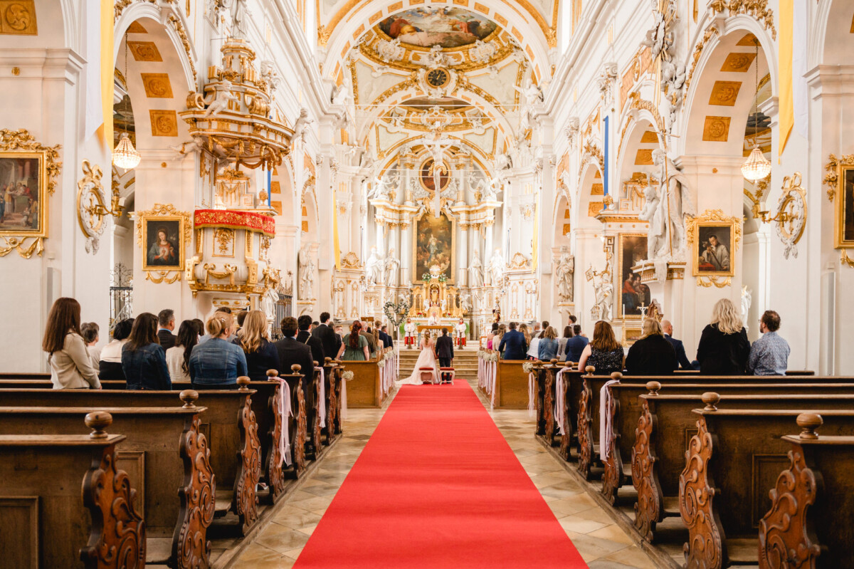Innenansicht einer großen Kirche. Menschen sitzen in Kirchenbänken zu beiden Seiten eines roten Teppichs, der zu einem kunstvollen Altar führt. Die Decke ist reich verziert, und die Wände sind mit Kunstwerken geschmückt.