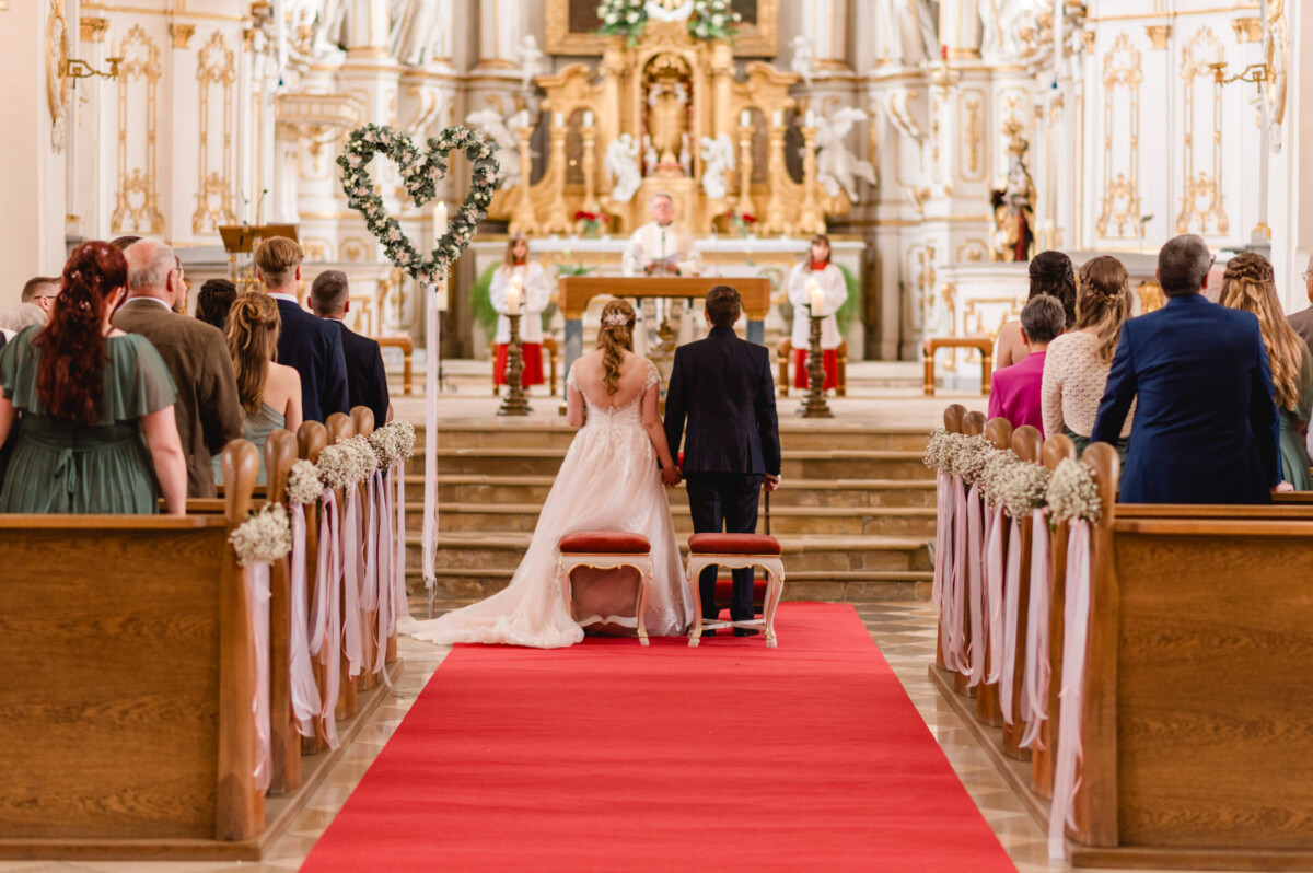 christian-lorenz.net Ein Paar kniet während einer Hochzeitszeremonie in einer reich verzierten Kirche vor dem Altar, während die Gäste auf beiden Seiten eines mit Blumen und Bändern geschmückten roten Teppichs auf dem Gang sitzen.
