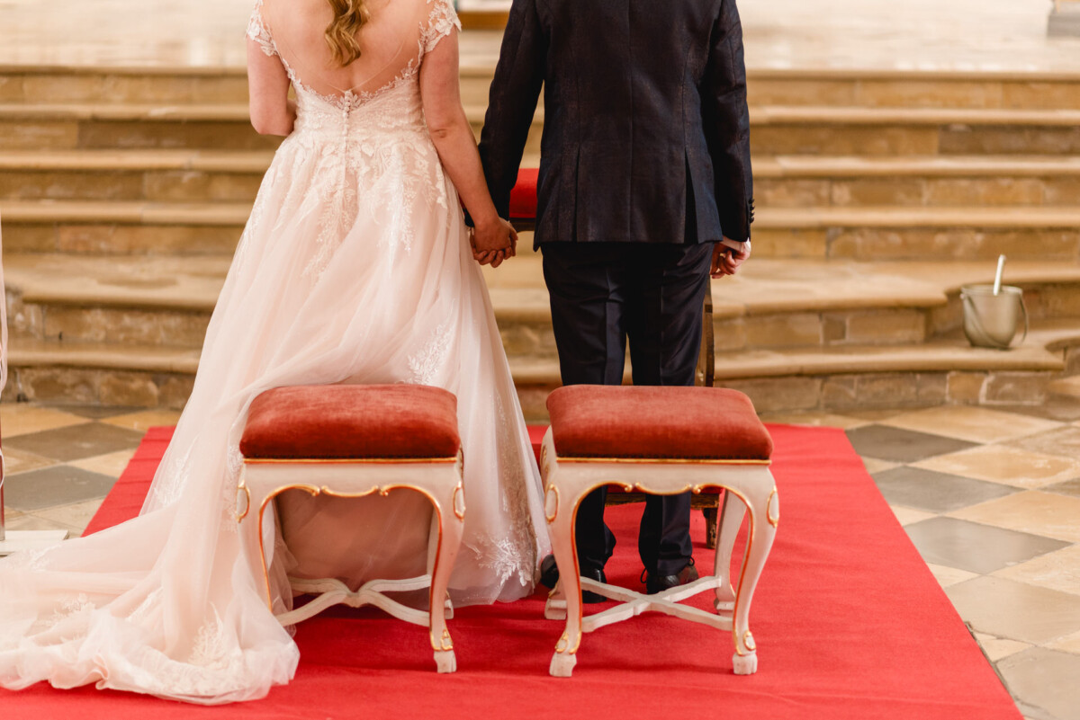 christian-lorenz.net Braut und Bräutigam stehen Hand in Hand vor zwei roten Hockern auf einem roten Teppich und blicken auf eine Reihe Steinstufen.
