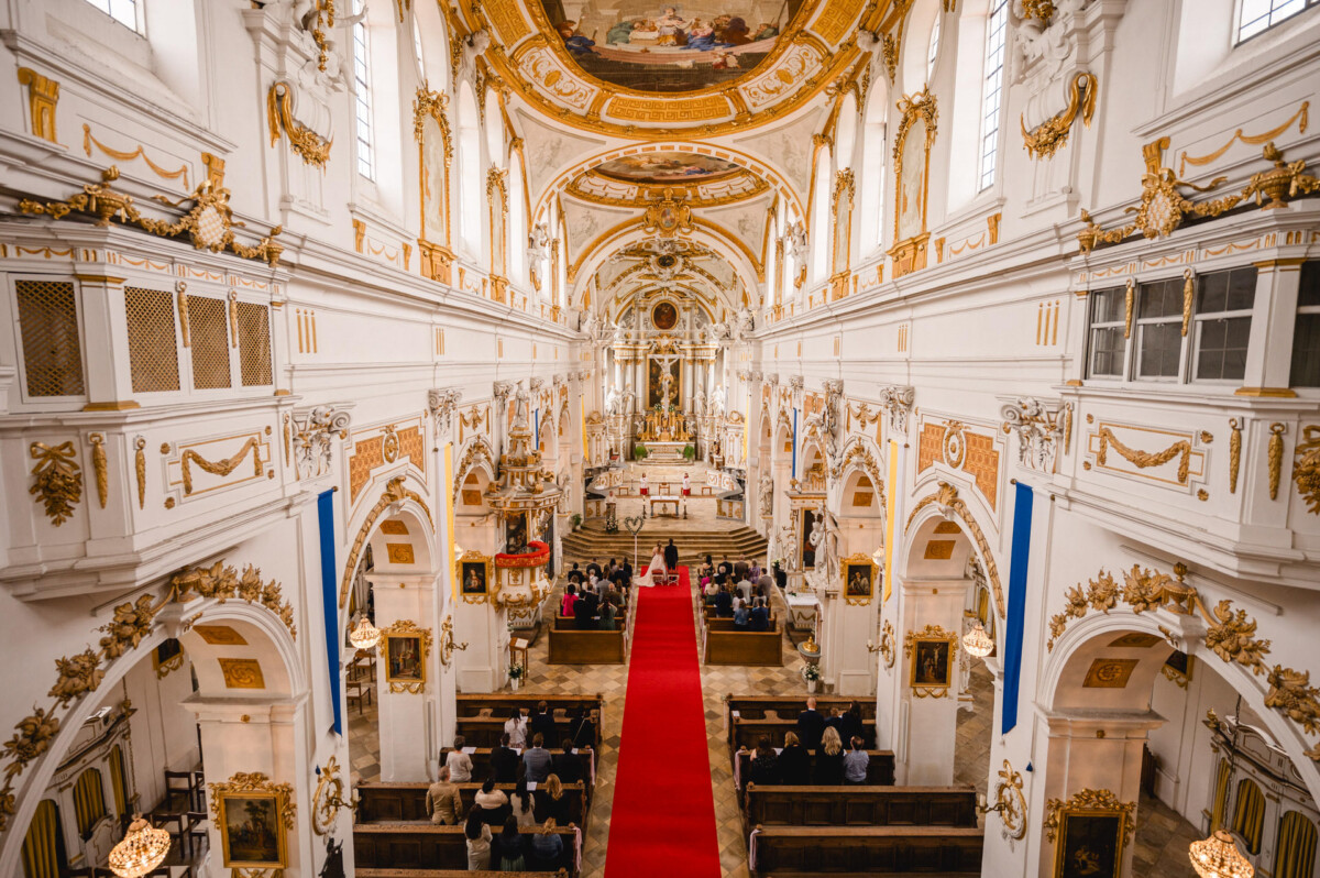 Innenraum einer kunstvollen Kirche mit goldenen Verzierungen und hohen Decken. Ein roter Teppich führt zum Altar, wo zu beiden Seiten eine Gruppe von Menschen sitzt.