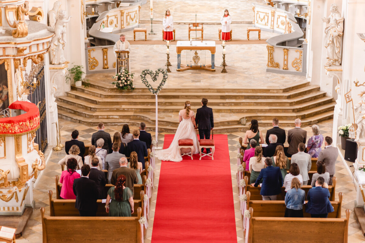 christian-lorenz.net Ein Paar steht bei einer kirchlichen Trauung am Altar. Die Gäste sitzen auf Holzbänken, ein roter Teppich führt nach vorne. Drei Personen stehen vor dem Altar.