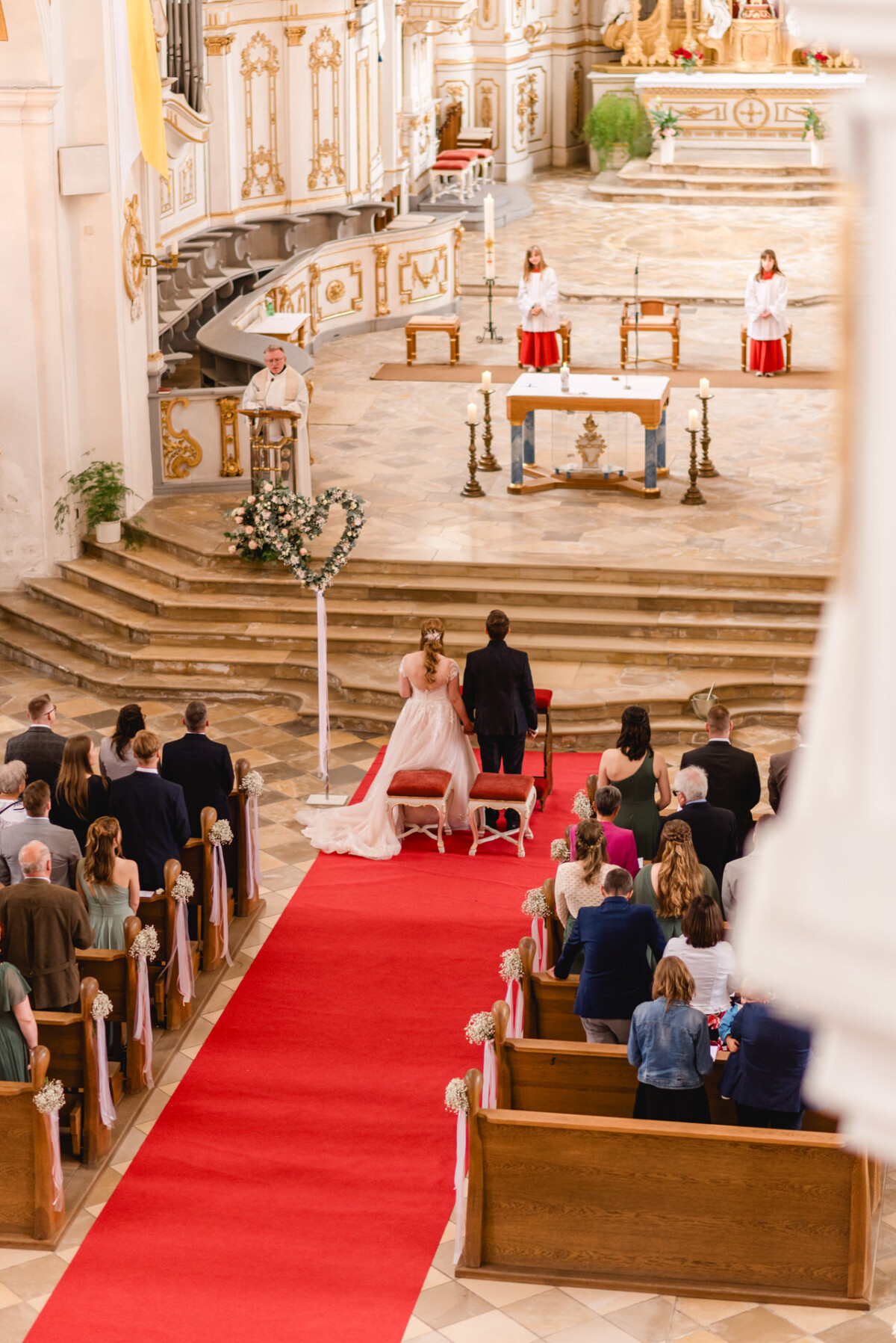 christian-lorenz.net Ein Paar sitzt während einer Hochzeitszeremonie vor dem Altar in einer Kirche. Die Gäste sitzen in den Kirchenbänken, und die Geistlichen in Weiß stehen am Altar. Ein roter Teppich führt den Gang entlang.