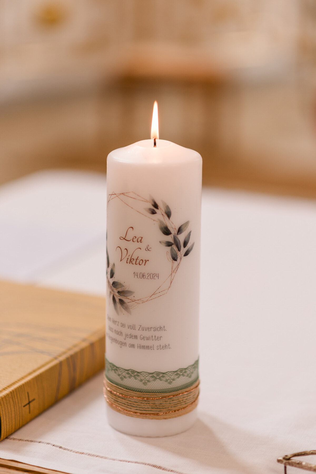 christian-lorenz.net Eine brennende weiße Kerze mit der Aufschrift „Lea & Viktor 14.06.2024“, umgeben von einem Blumenmuster, steht auf einem mit Stoff bedeckten Tisch.