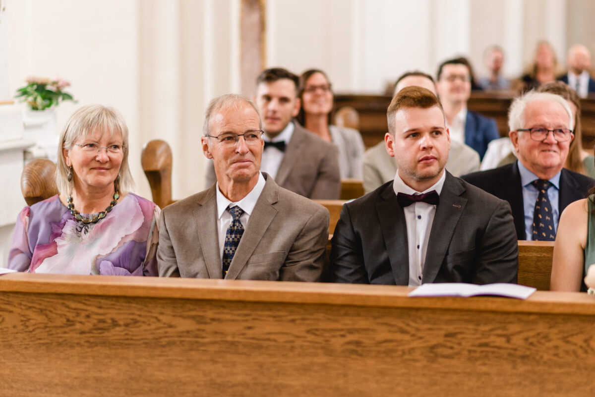 christian-lorenz.net Drei Personen sitzen in der ersten Reihe einer Kirche, sind festlich gekleidet und beobachten eine Veranstaltung. Andere sitzen hinter ihnen.