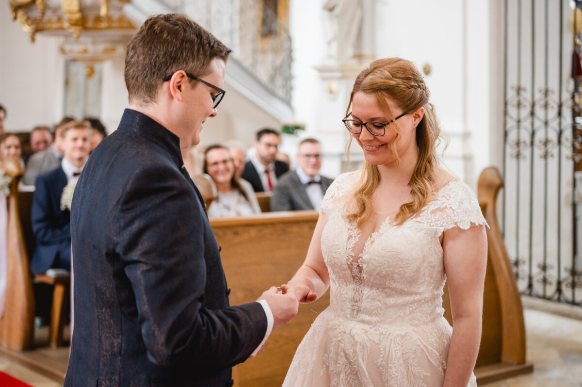 Ein Paar tauscht während einer Hochzeitszeremonie in einer Kirche die Ringe aus, während im Hintergrund sitzende Gäste zuschauen.