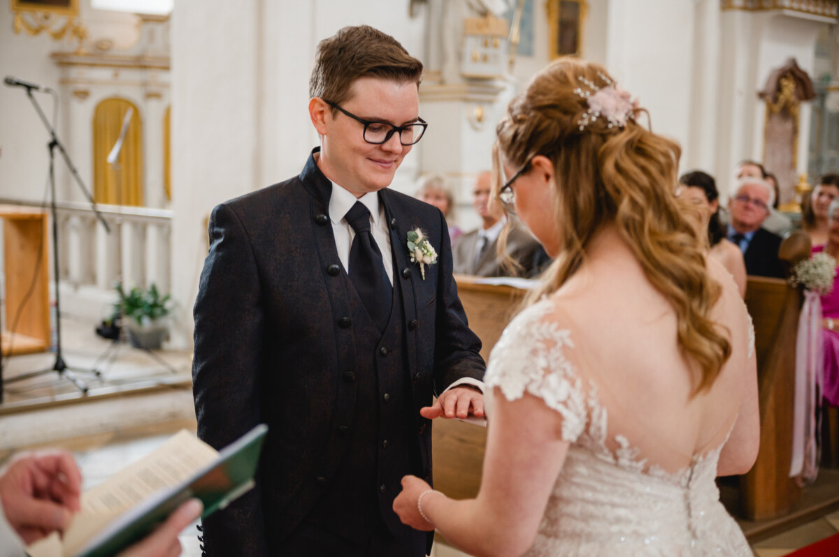 Ein Paar tauscht während einer Hochzeitszeremonie in einer Kirche, umgeben von Gästen, Ringe aus.