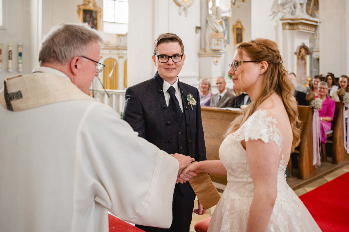 Braut und Bräutigam stehen in einer Kirche händchenhaltend am Altar. Ein Geistlicher führt den Gottesdienst durch. Im Hintergrund sitzen Gäste.