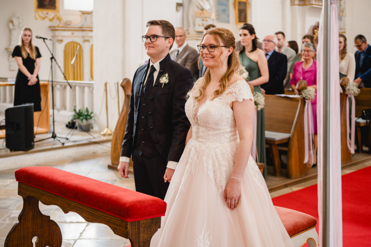 Während einer Hochzeitszeremonie in einer Kirche stehen Braut und Bräutigam am Altar, während die Gäste in den Kirchenbänken hinter ihnen sitzen.