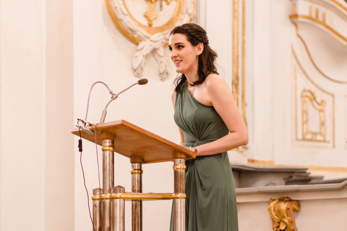 christian-lorenz.net Eine Frau in einem grünen Kleid spricht an einem Podium mit einem Mikrofon in einem reich verzierten Raum mit goldenen Akzenten.