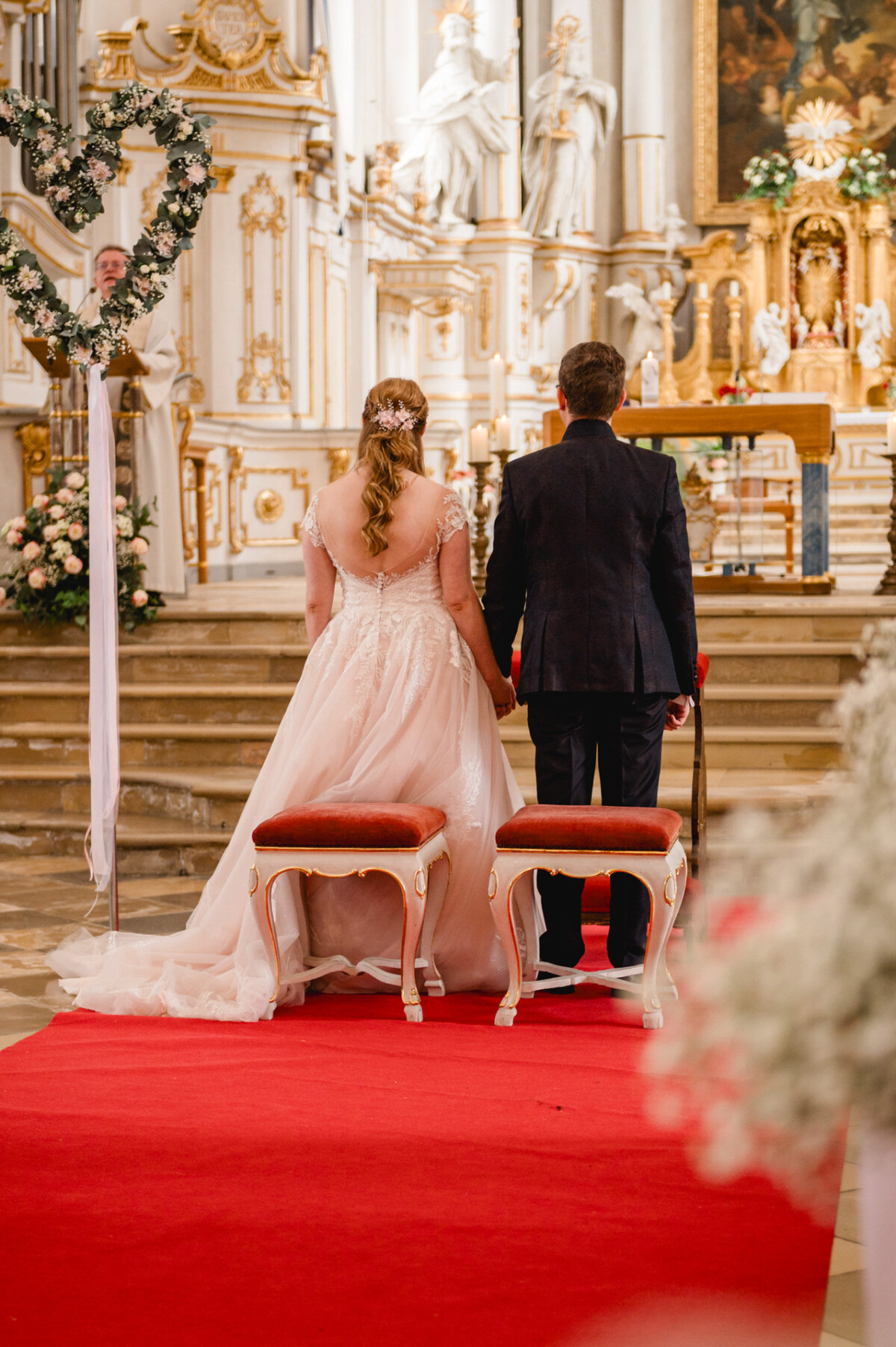 Ein Brautpaar steht mit abgewandtem Gesicht auf einem roten Teppich in einer geschmückten Kirche am Altar.