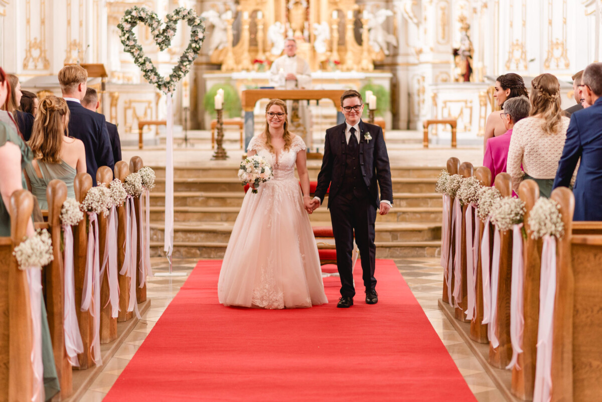 christian-lorenz.net Braut und Bräutigam schreiten nach ihrer Hochzeitszeremonie in einer Kirche, umgeben von Gästen, den Gang entlang.