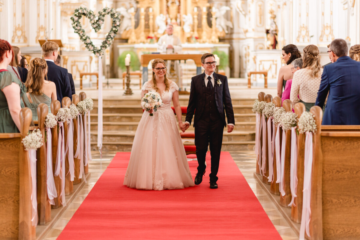 christian-lorenz.net Ein Brautpaar geht in einer geschmückten Kirche Hand in Hand den Gang entlang, während die Gäste auf mit Blumen geschmückten Holzbänken sitzen.