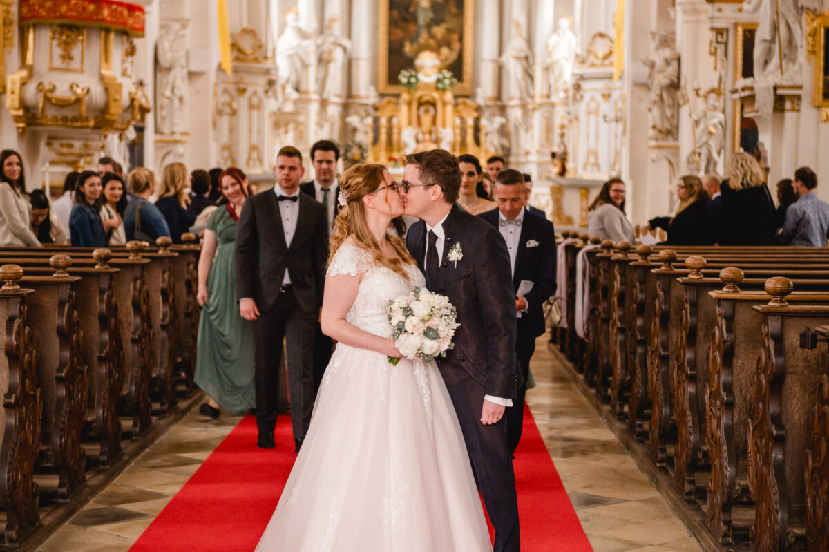 Ein Brautpaar küsst sich, während es umgeben von Gästen auf einem roten Teppich in einer Kirche den Gang entlanggeht.