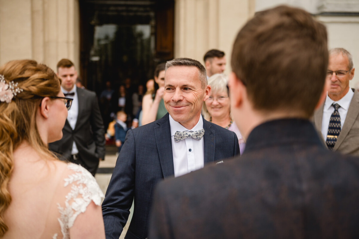 Ein Mann im Anzug lächelt während einer geselligen Runde vor einem Gebäude, umgeben von Menschen, darunter eine Frau in einem weißen Kleid.