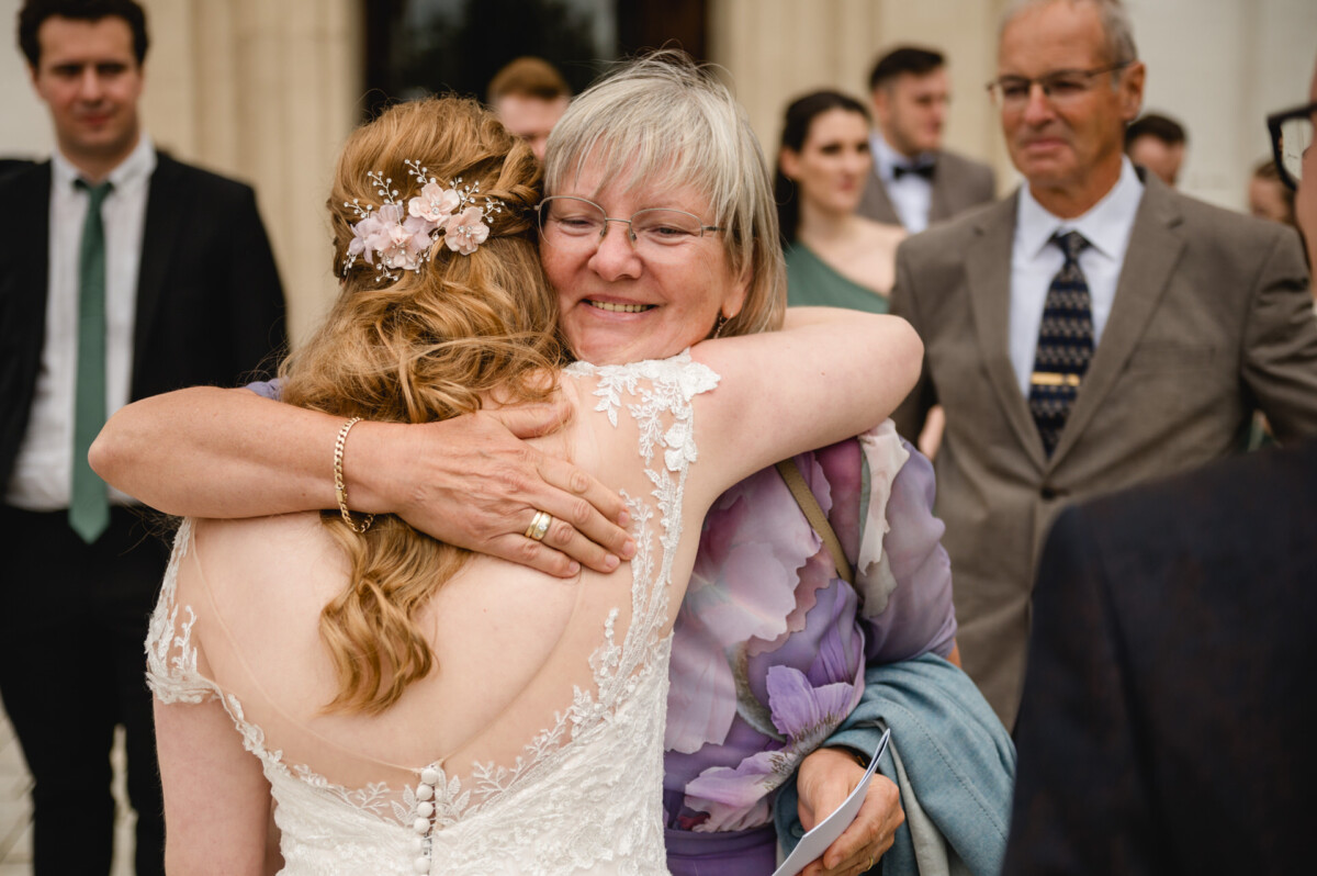 Eine Frau im Spitzenkleid umarmt eine andere Frau mit Brille und geblümtem Outfit. Im Hintergrund sind Menschen in Abendgarderobe zu sehen.