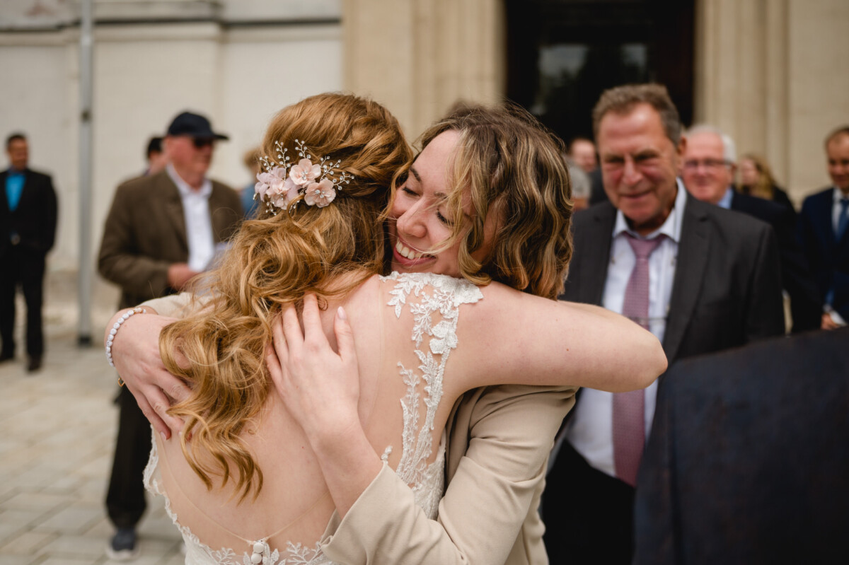 Zwei Frauen umarmen sich bei einer Veranstaltung im Freien, im Hintergrund sind weitere in Abendgarderobe zu sehen.