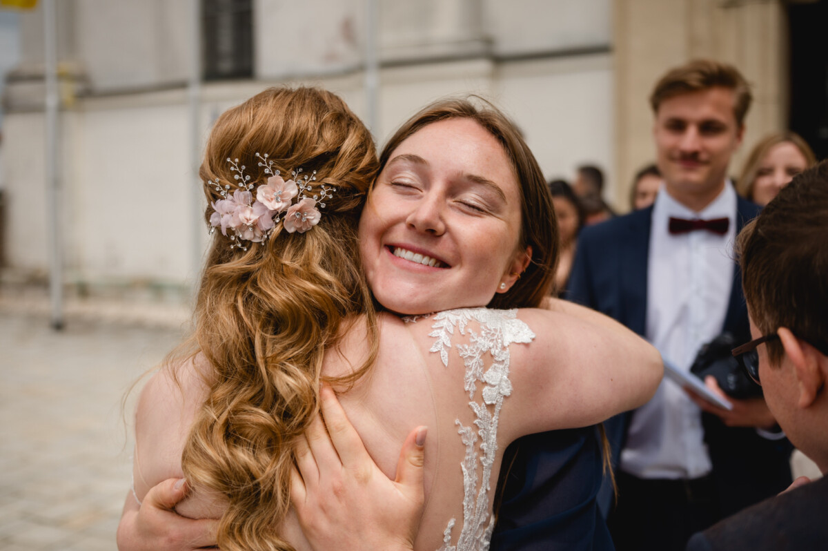 Zwei Frauen umarmen sich freudig; eine trägt ein weißes Kleid mit Spitzendetails und einem floralen Haarschmuck. Andere stehen im Hintergrund, darunter ein Mann im Anzug mit Fliege.