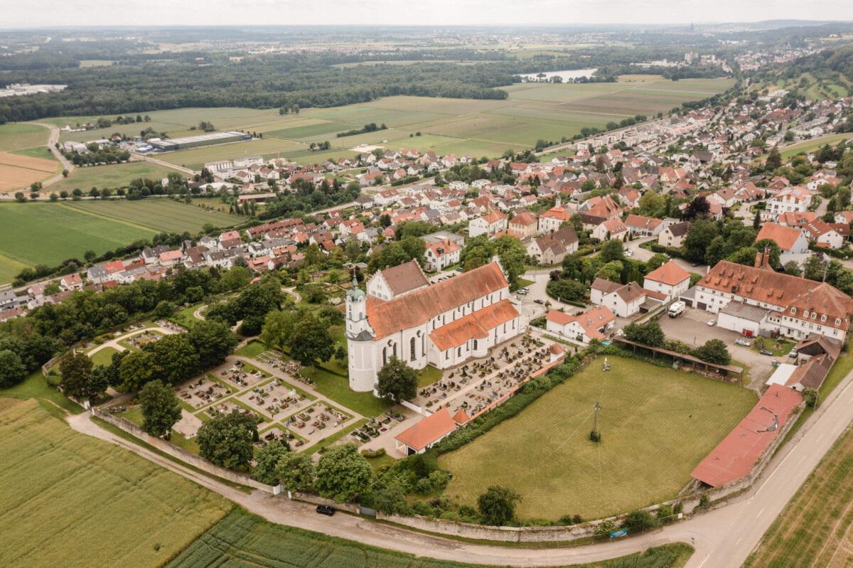 Luftaufnahme einer Stadt mit einer großen Kirche im Zentrum, umgeben von Wohnhäusern, Feldern und einem Friedhof.