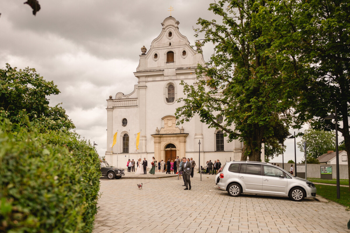 Menschen versammeln sich vor einer weißen historischen Kirche mit barocker Fassade. Autos parken in der Nähe, Bäume säumen den Weg. Über ihnen ist ein bewölkter Himmel.