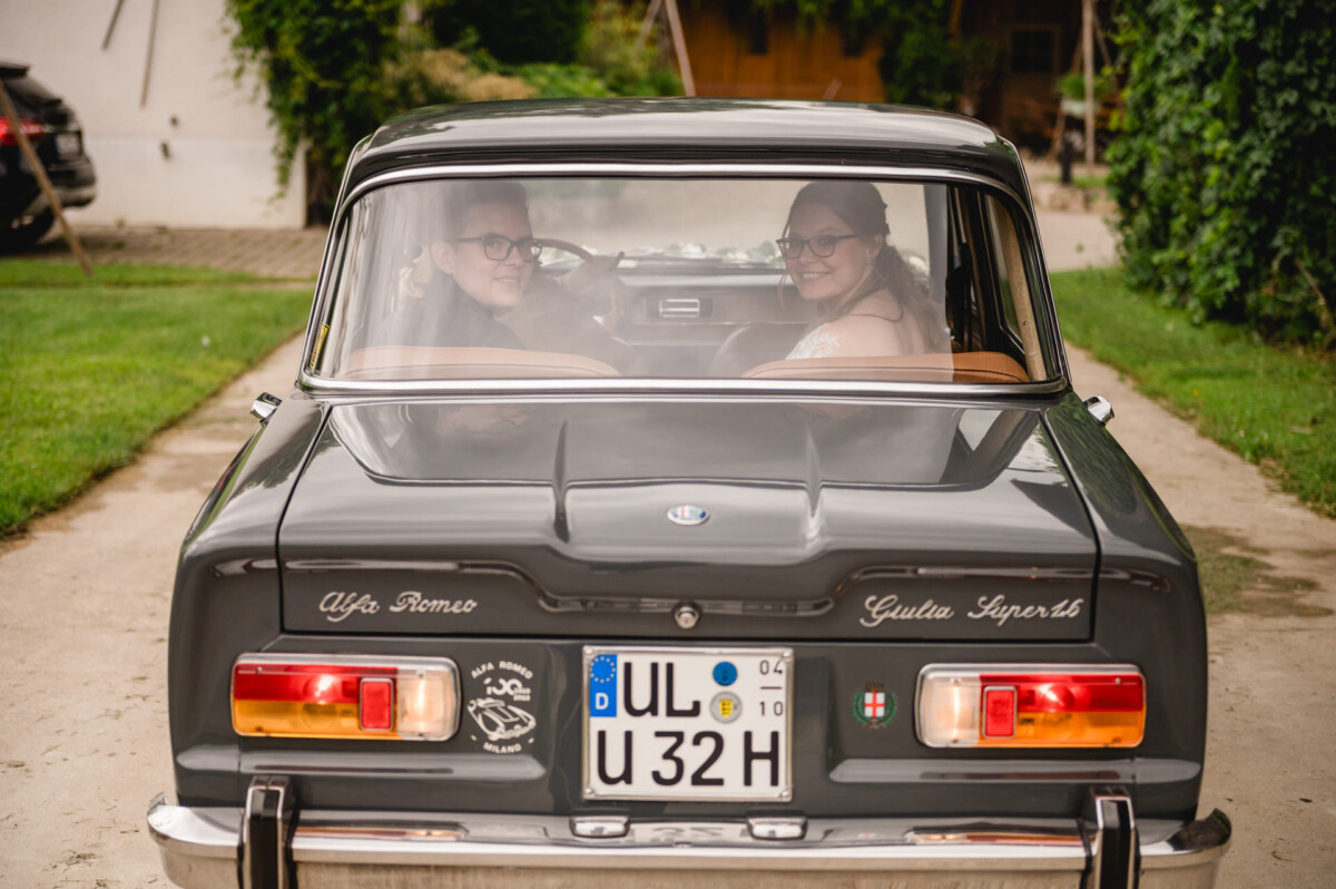 Ein Paar sitzt von hinten in einem alten Alfa Romeo Giulia Super. Das Auto ist auf einer von Grün gesäumten Auffahrt geparkt.