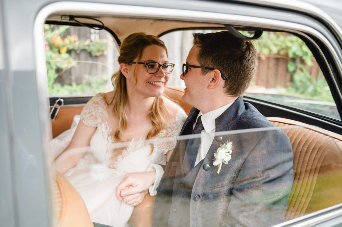 Braut und Bräutigam sitzen in einem Auto, halten Händchen und lächeln sich an, vor dem Fenster ist Grün zu sehen.
