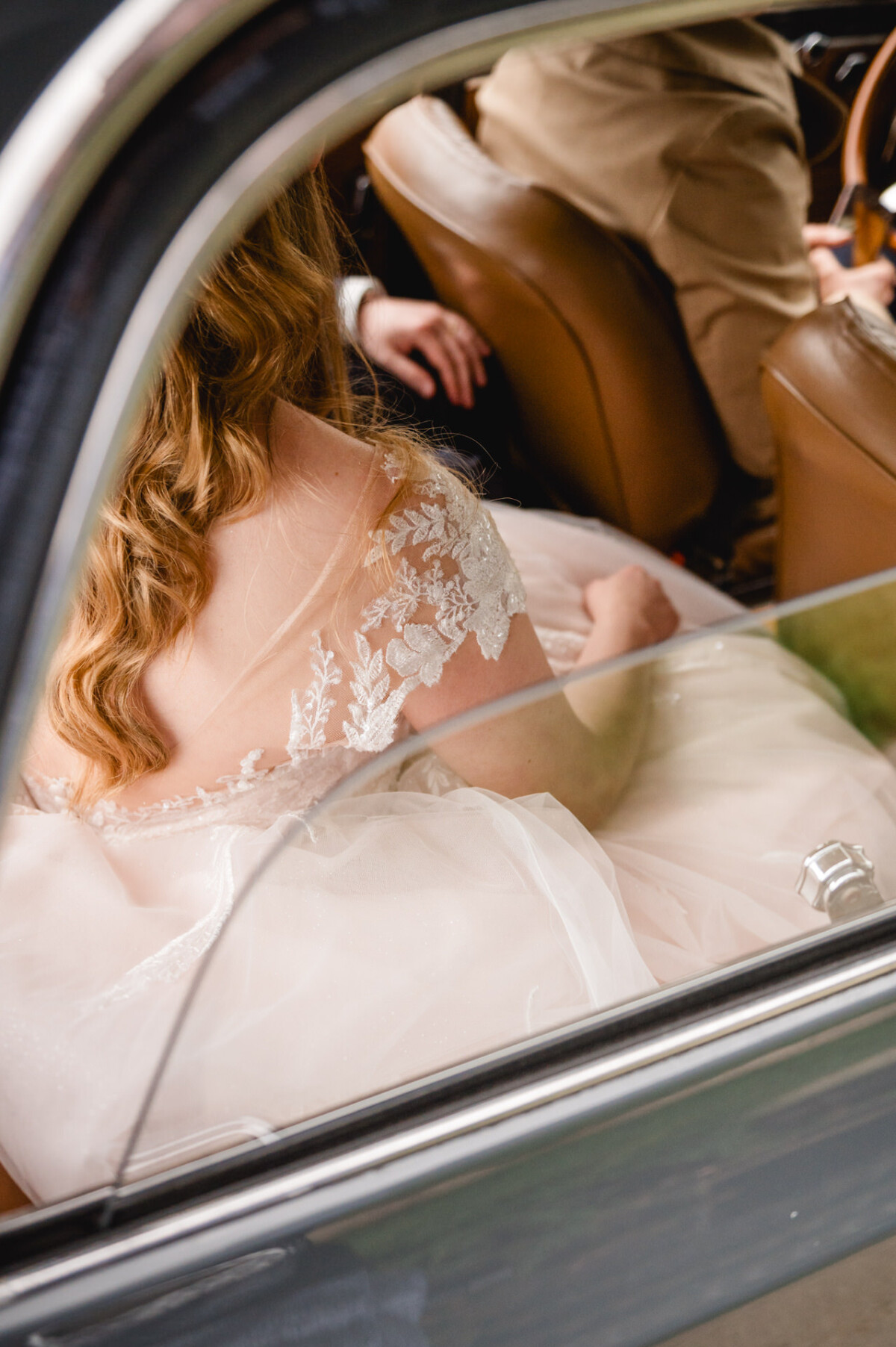 Auf dem Rücksitz eines Autos sitzt eine Person in einem weißen Spitzenkleid. Auf dem Vordersitz ist eine weitere Person in einem hellbraunen Anzug zu sehen.