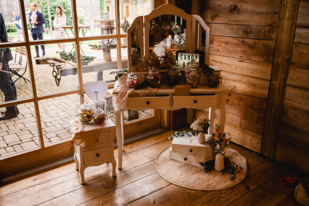 Candy-Bar-Aufbau auf einem hölzernen Frisiertisch mit Bonbongläsern, Blumen und einem „Candy Bar“-Schild in einem rustikalen Holzraum mit großen Fenstern im Hintergrund.