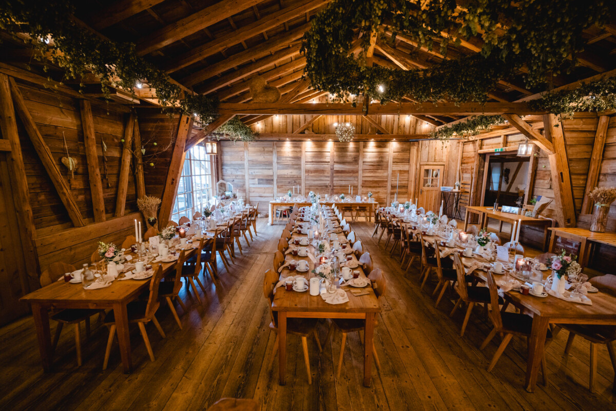 Rustikaler Speisesaal mit Holzdekor, gedeckt für ein Bankett mit in Reihen angeordneten Tischen, geschmückt mit weißen Tischdecken, Blumen und Geschirr. Warme Beleuchtung schafft eine gemütliche Atmosphäre.