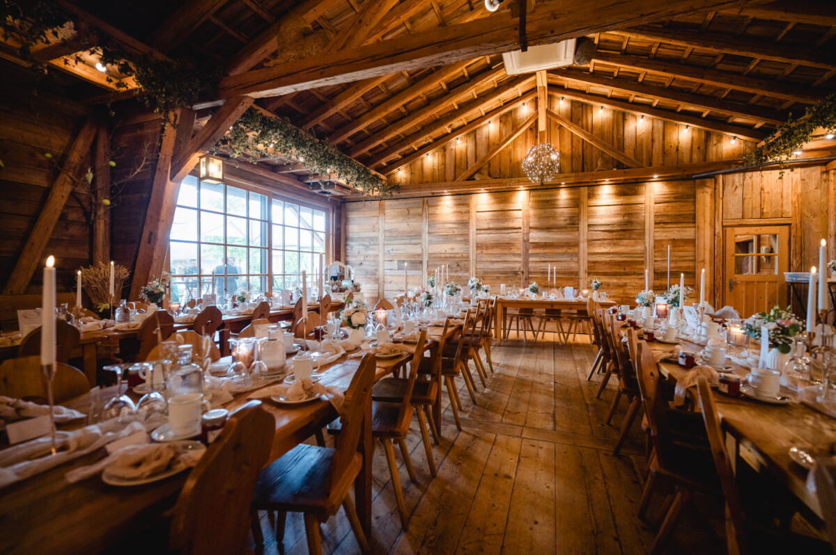 Rustikaler Bankettsaal aus Holz mit langen Tischen, dekoriert mit Kerzen und Grünpflanzen. Warmes Licht unterstreicht die gemütliche Atmosphäre.